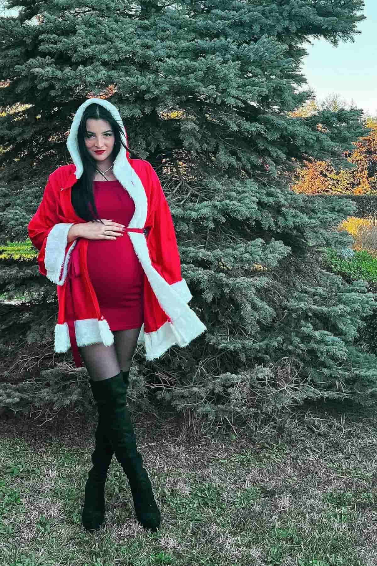 A woman in a red and white Santa-style outfit stands outdoors in front of a large evergreen tree, with one hand on her belly.