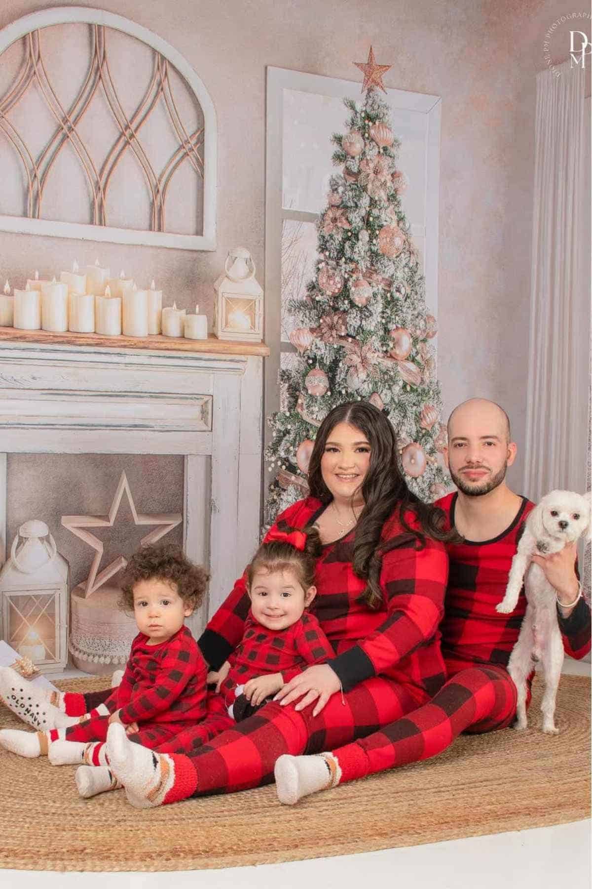 A family of four wearing matching red plaid pajamas sits by a Christmas tree and fireplace, with a small white dog held by the man.