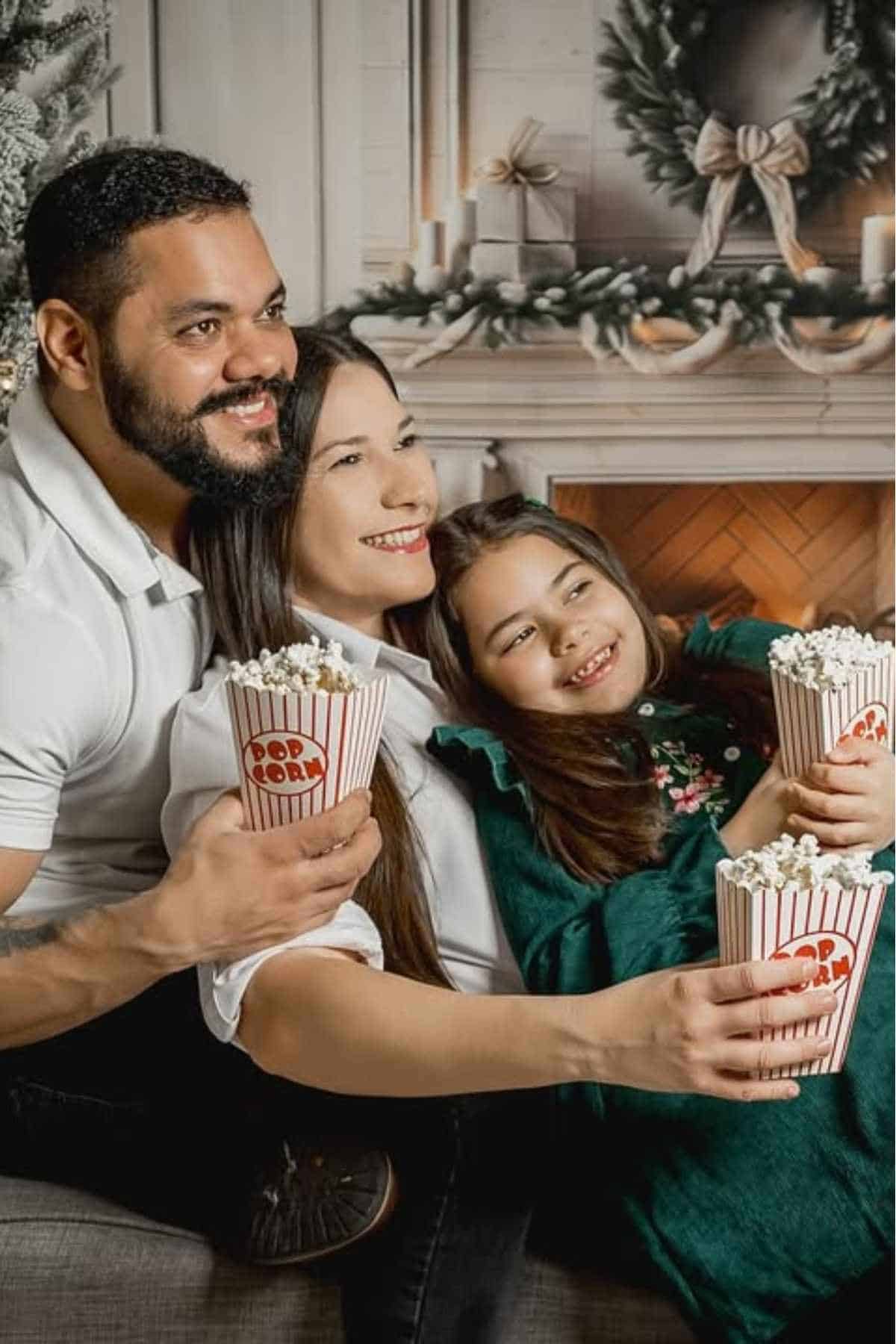A man, woman, and child sit together holding popcorn, smiling, with a decorated fireplace and Christmas tree in the background.