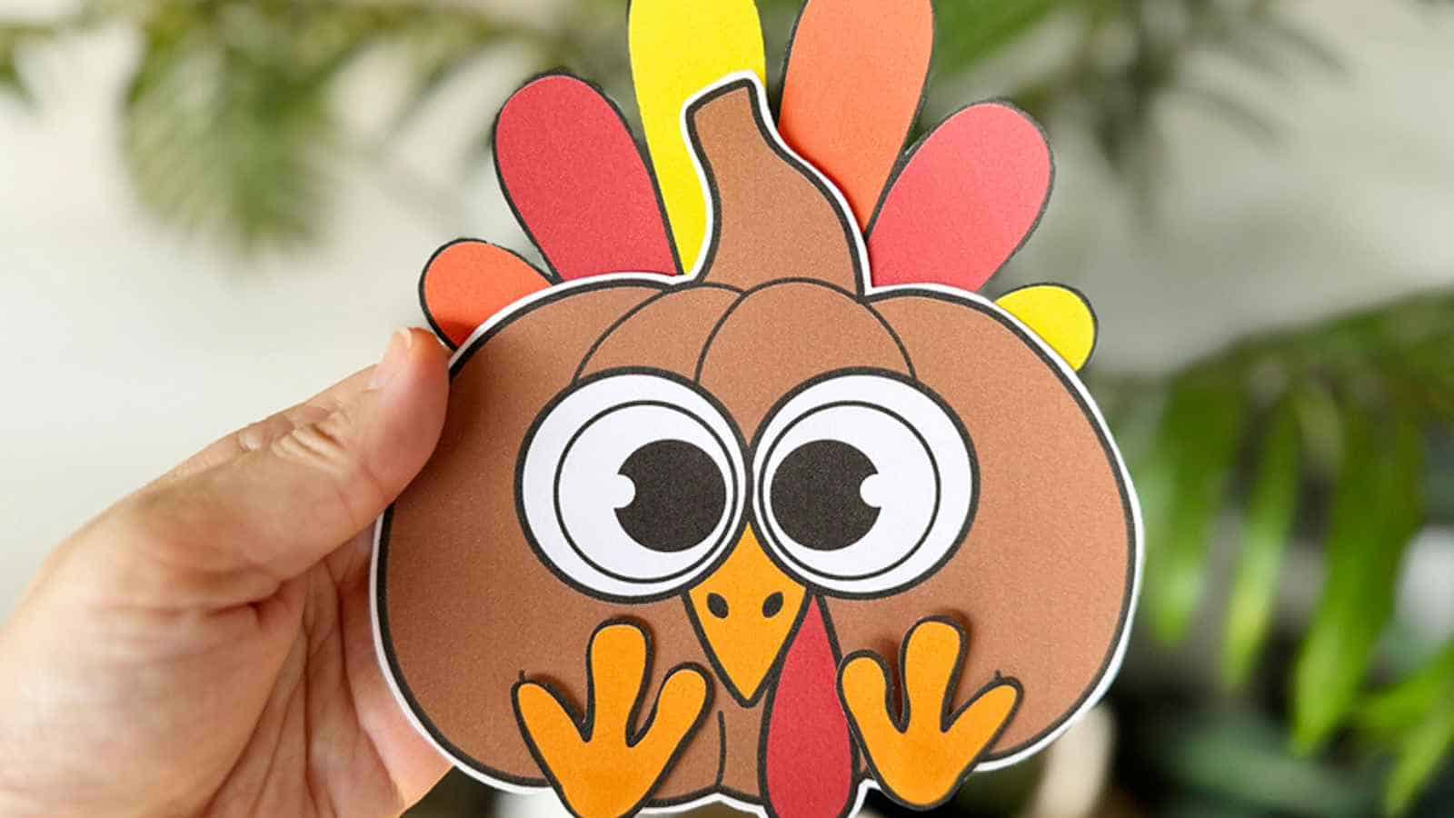A hand holds a colorful paper craft featuring a cartoon turkey with large eyes and orange, red, and yellow feathers.