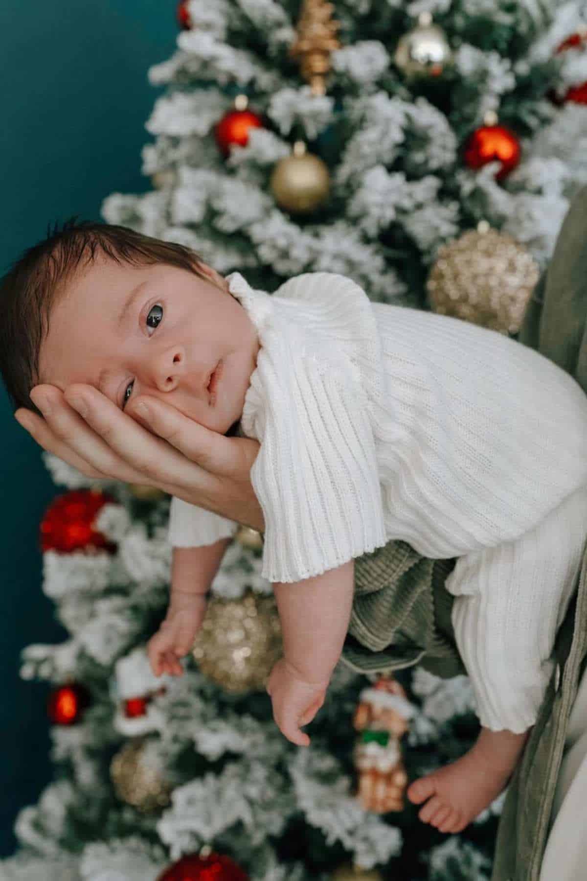 A baby dressed in white is cradled in an adult's hand in front of a decorated Christmas tree with red and gold ornaments.