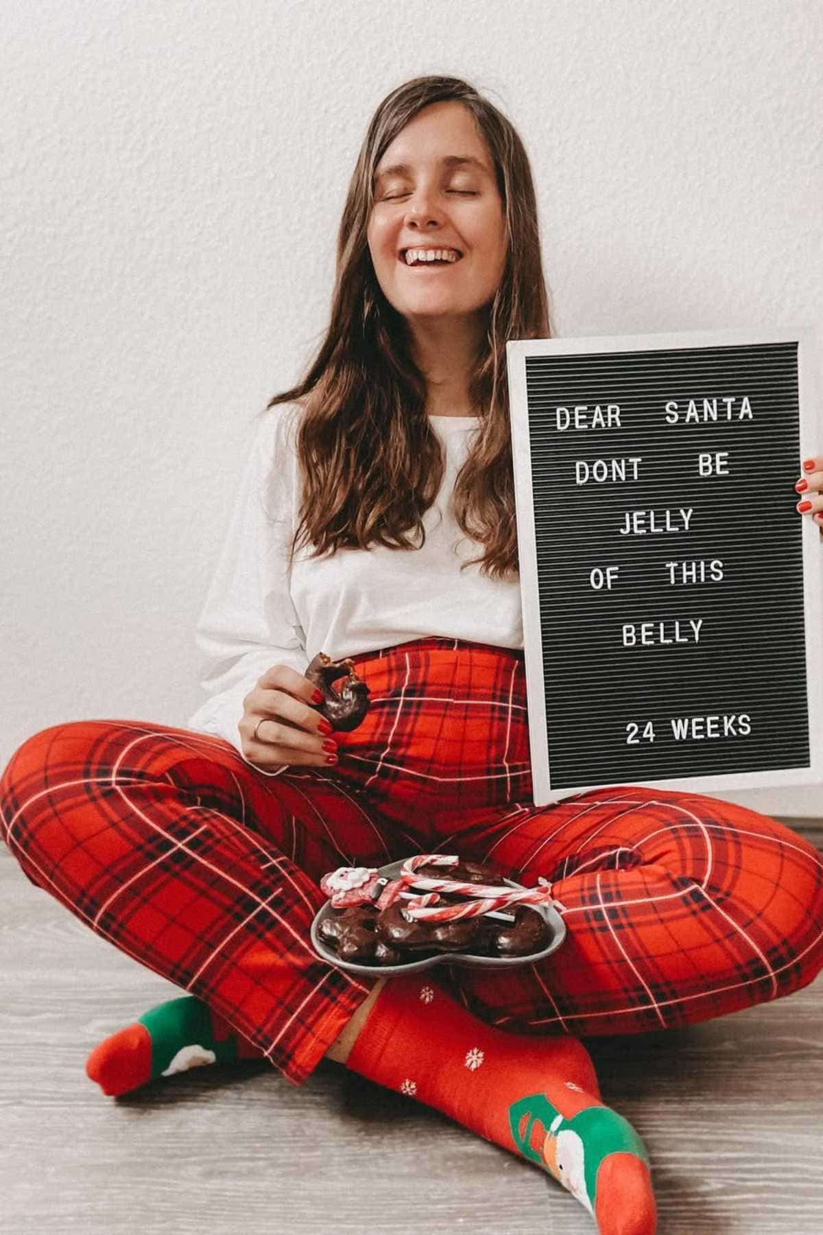Woman in red plaid pants and festive socks sits on the floor, holding a doughnut and a letterboard that reads, "Dear Santa don't be jelly of this belly 24 weeks.