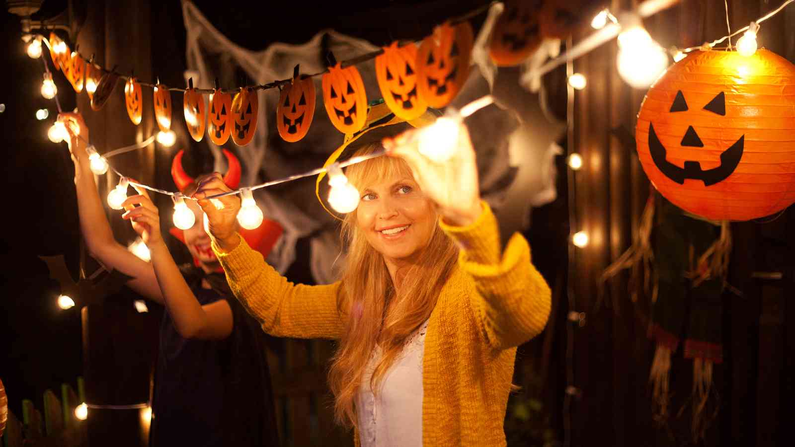 Two people hang string lights among pumpkin and Halloween decorations, with one person smiling at the camera.