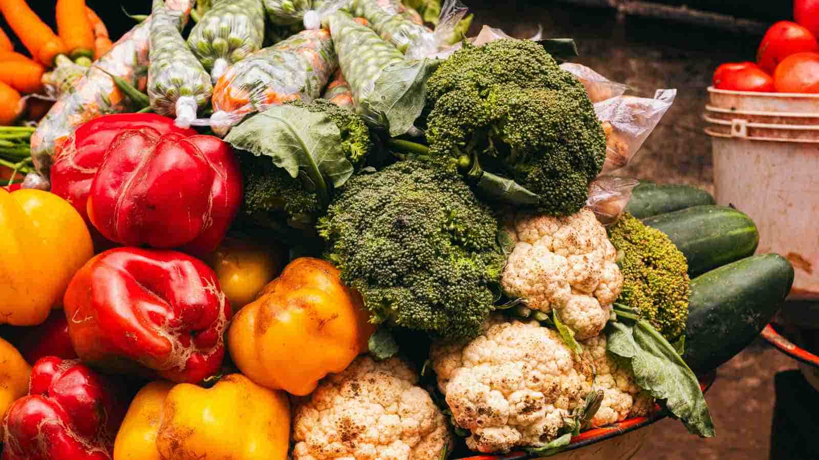 A display of fresh vegetables including red and yellow bell peppers, broccoli, cauliflower, zucchini, carrots, and tomatoes at a market stall.