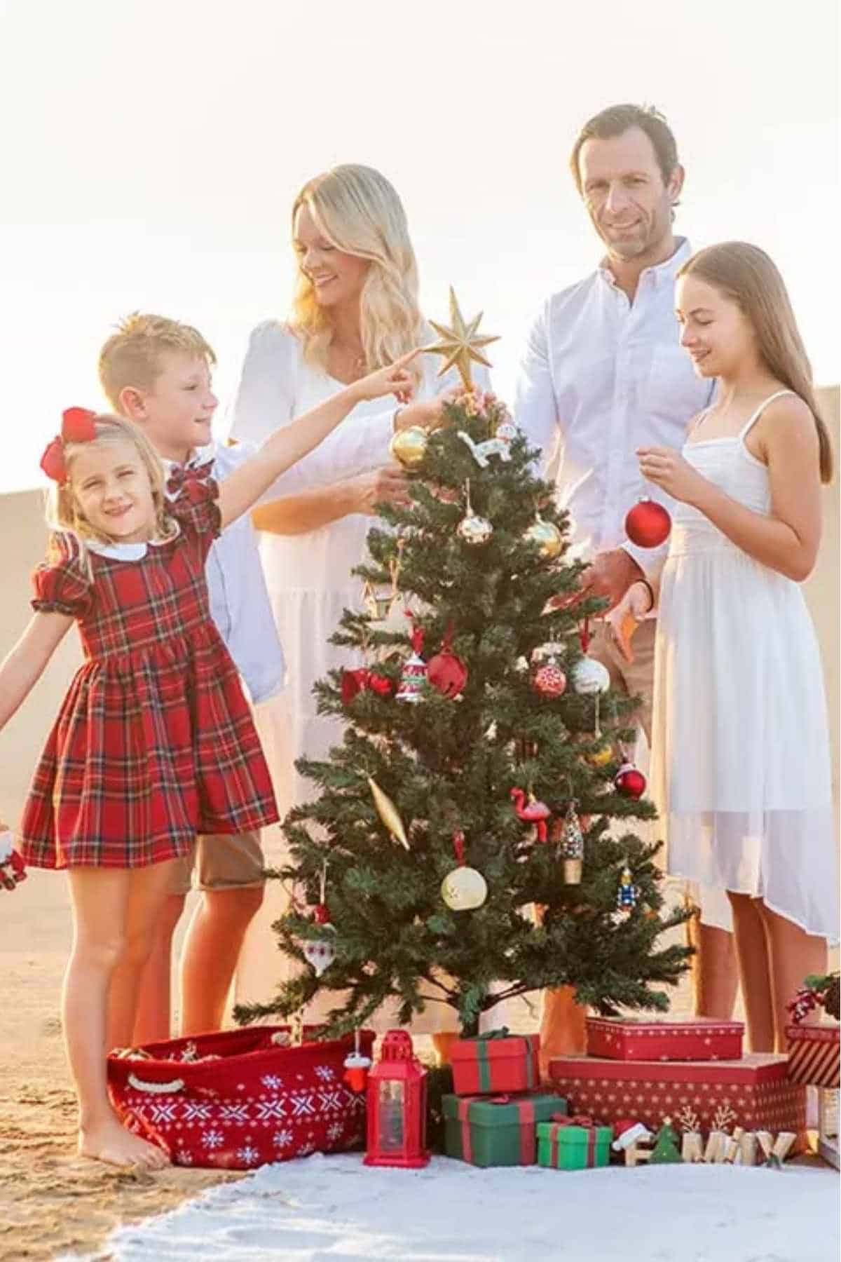 A family of five, dressed in white and red, decorates a small Christmas tree with ornaments and lights outdoors, surrounded by wrapped gifts.