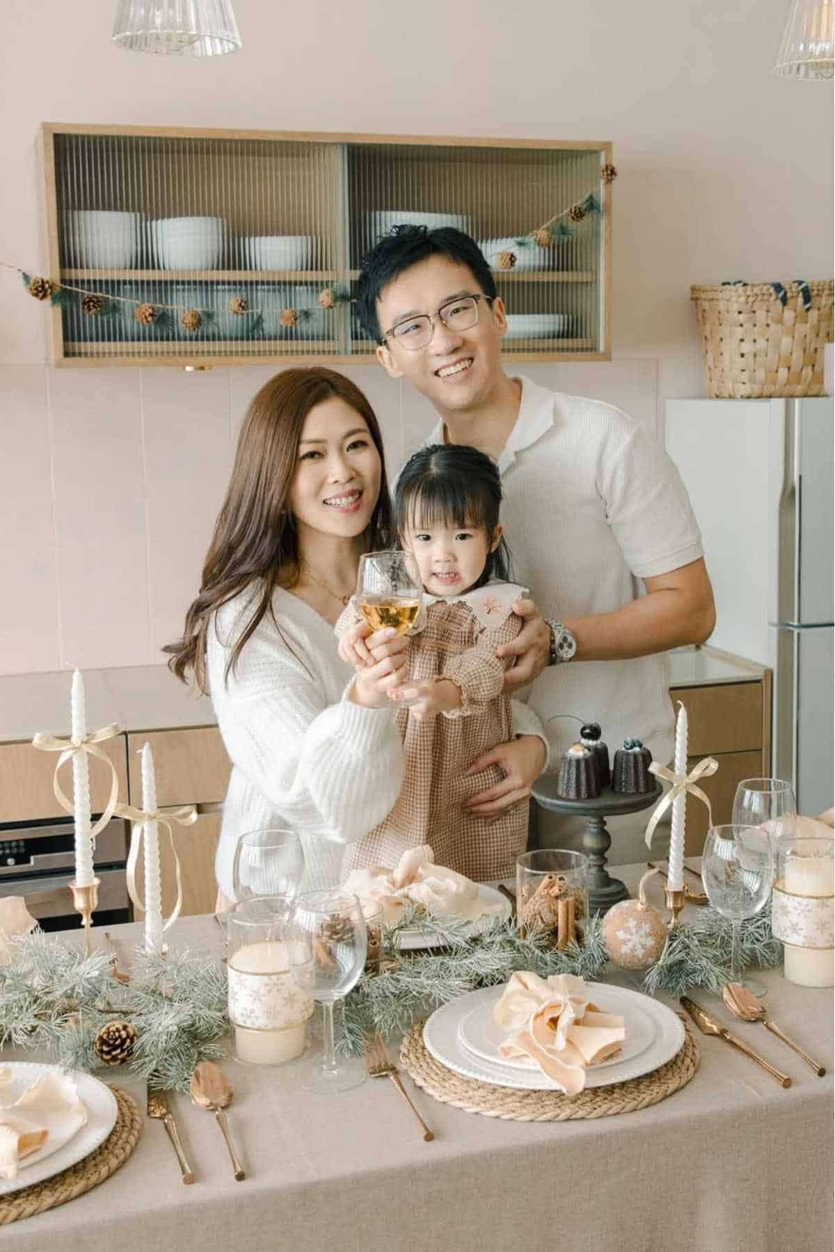 A family of three poses and smiles in a kitchen next to a decorated dining table set for a festive meal with candles, greenery, and tableware.