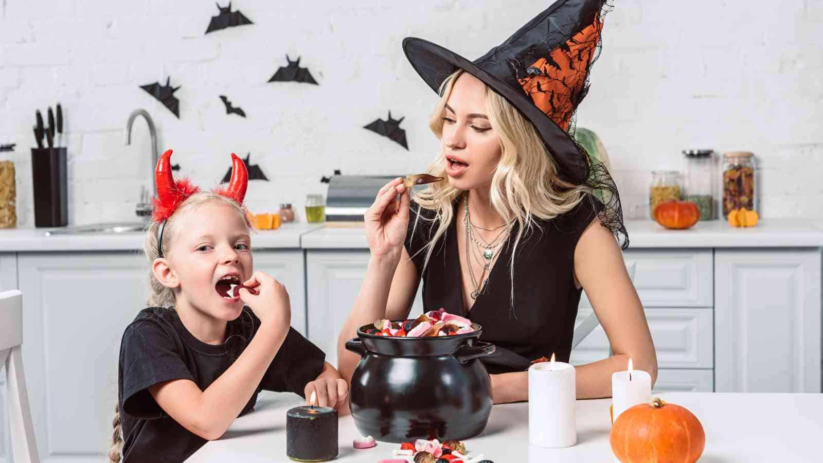 An adult and a child in Halloween costumes eat candy from a cauldron at a kitchen table decorated with pumpkins, candles, and paper bats.