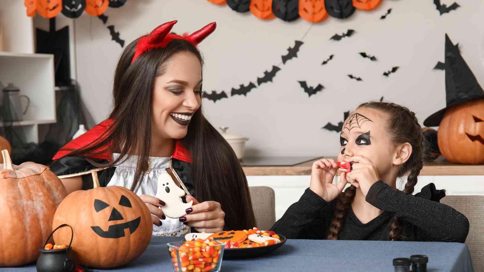 An adult and child in Halloween costumes sit at a table decorated with pumpkins and candy, smiling and eating treats. Halloween decorations are visible in the background.