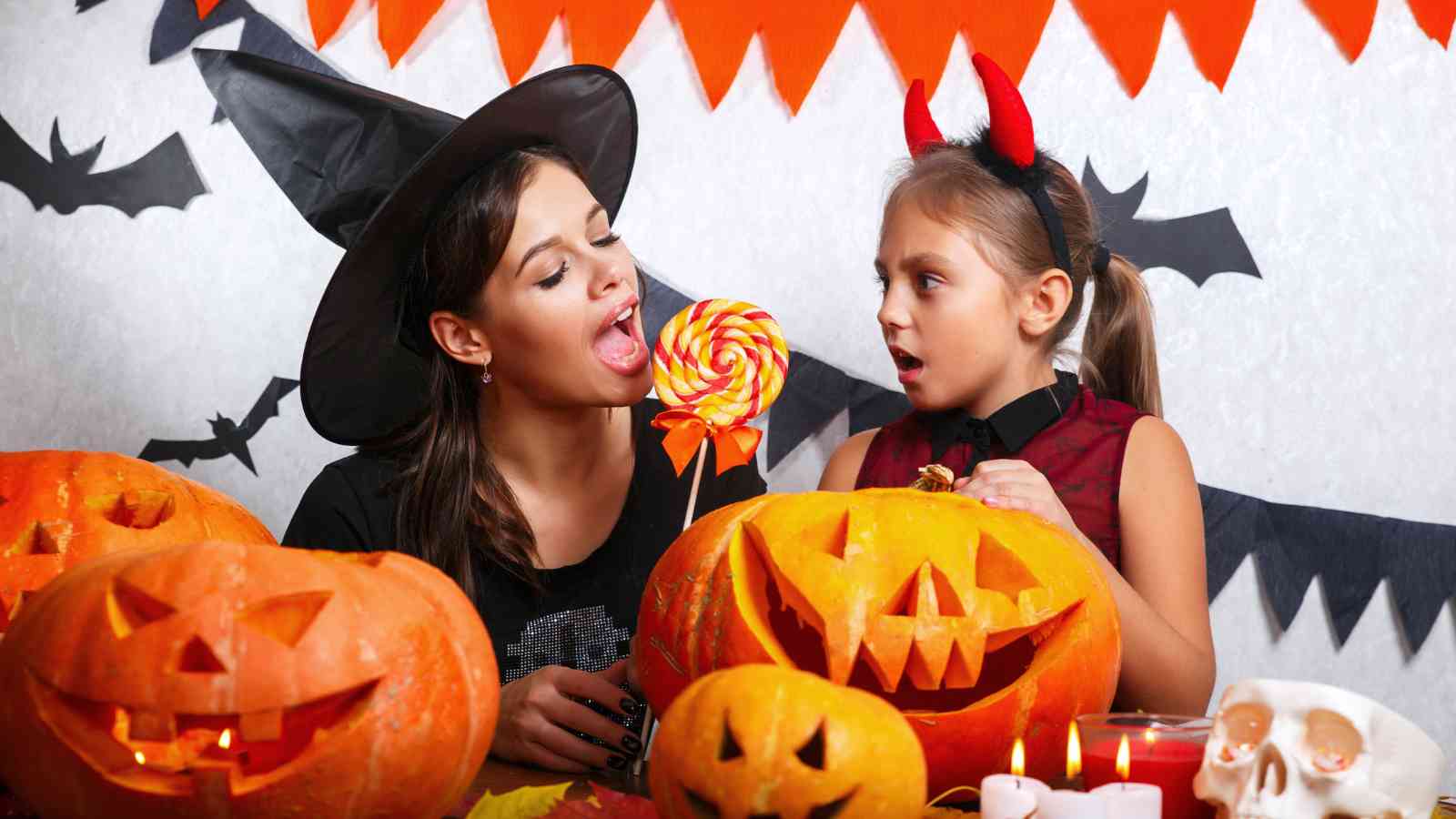 Two people in Halloween costumes sit at a table with carved pumpkins and candles, sharing a large lollipop, with festive decorations in the background.