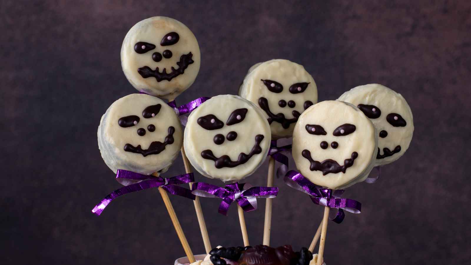 Six cookie pops decorated with white coating and black spooky faces, each tied with a purple ribbon, arranged against a dark background.