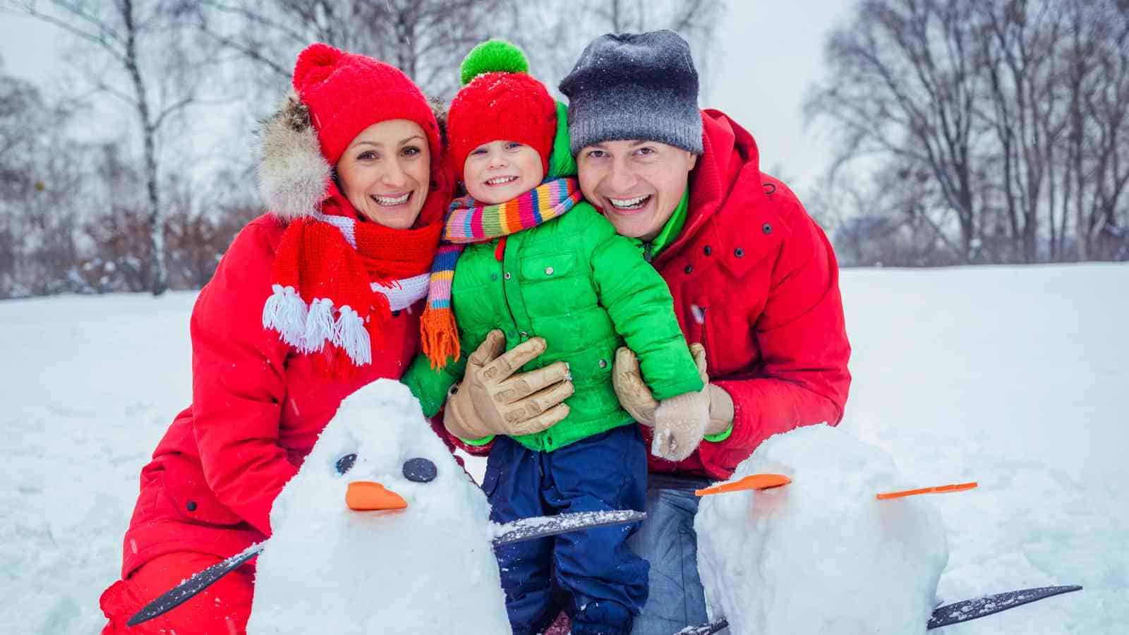 Three people in winter clothes kneel in the snow, smiling between two small snowmen with carrot noses. Leafless trees are visible in the background.