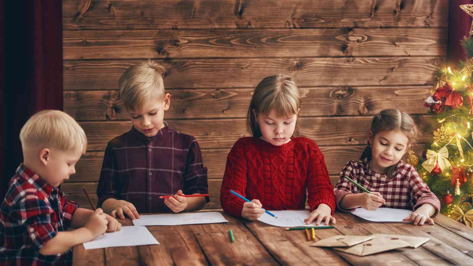Four children sit at a wooden table drawing on paper with colored pencils, with a decorated Christmas tree on the right side of the image.