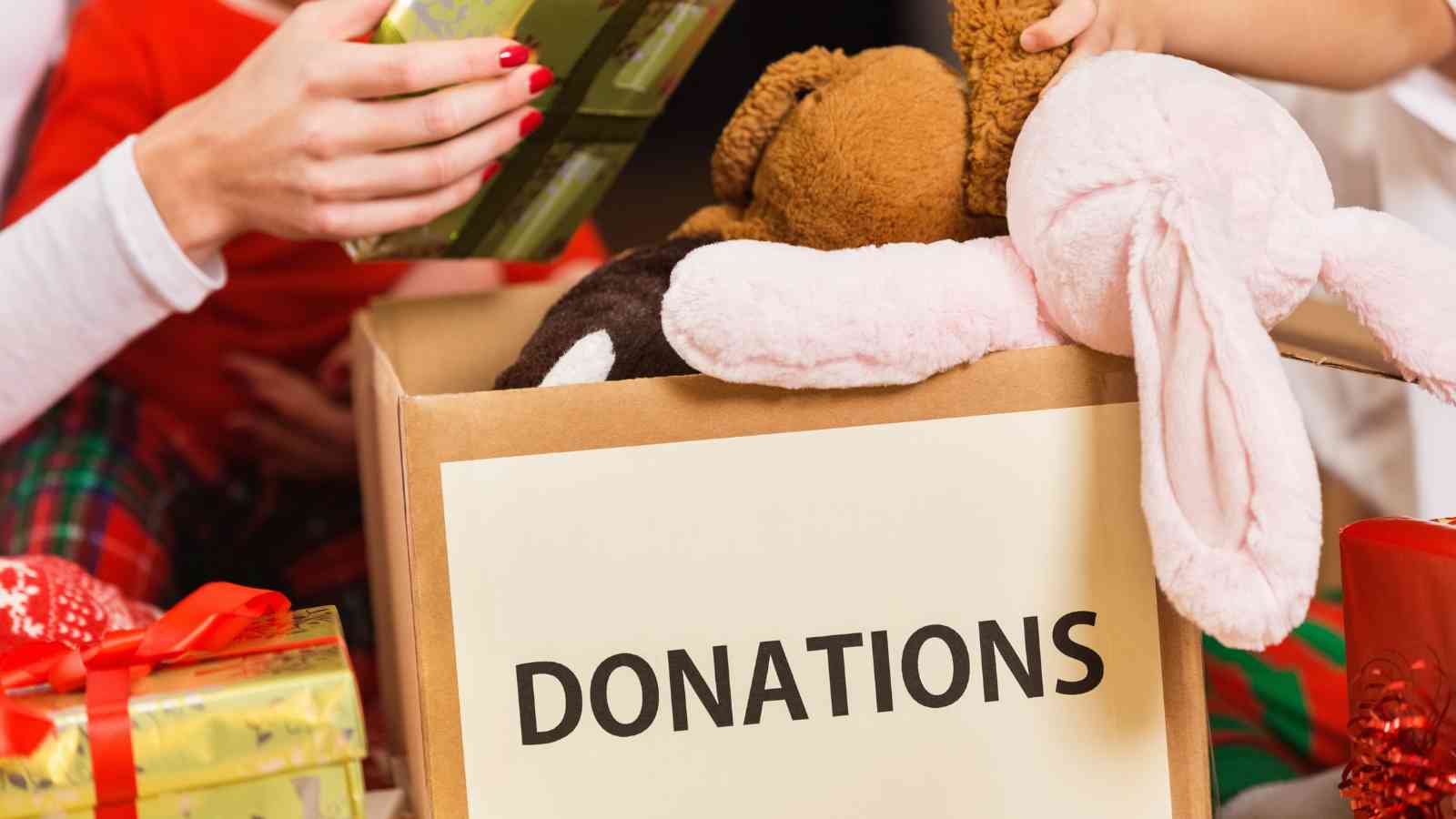 A cardboard box labeled "DONATIONS" filled with stuffed animals and toys, with hands placing more items inside.