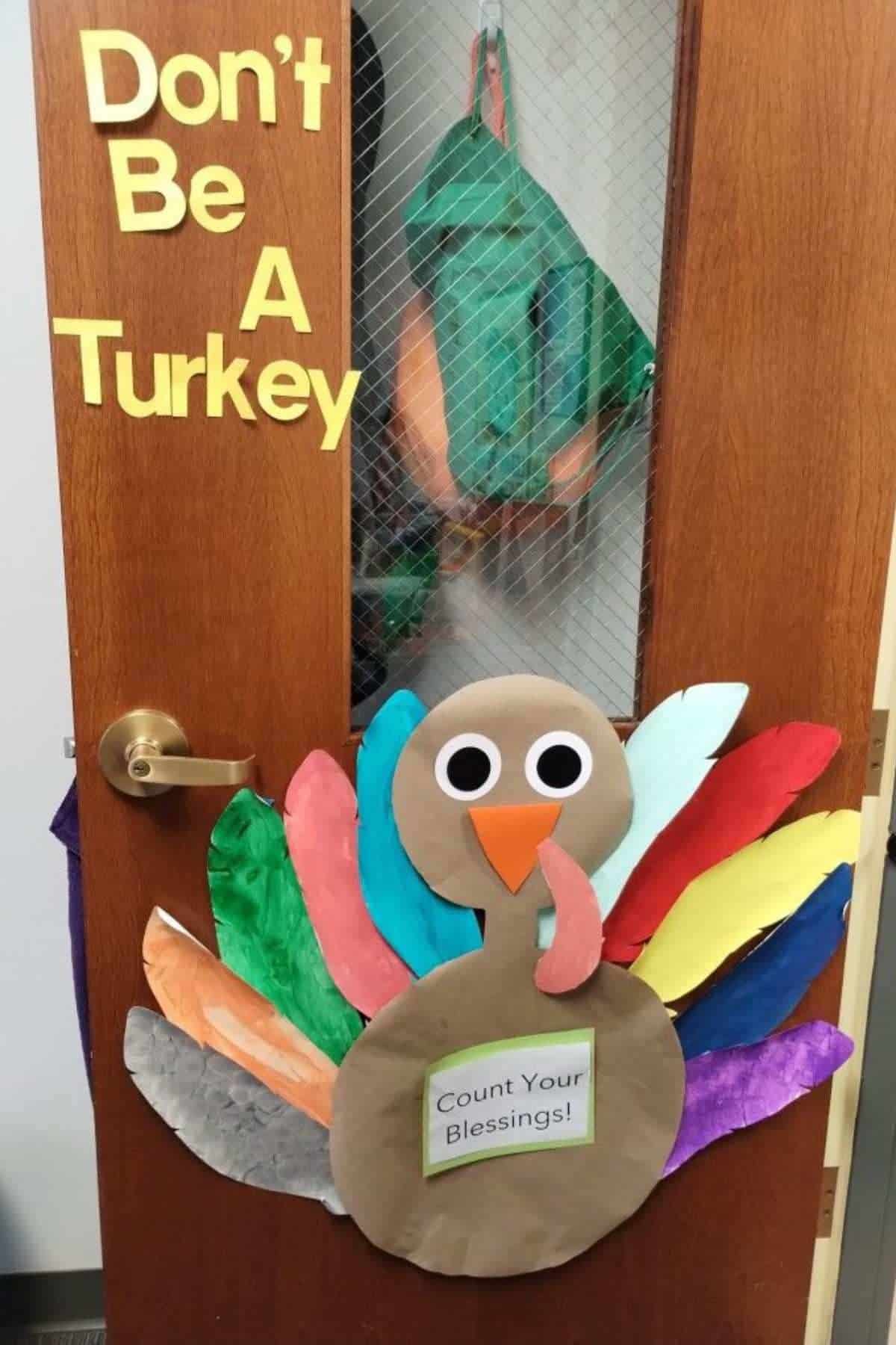 A door decorated with colorful paper turkey feathers, a large turkey face, and the words “Don’t Be A Turkey” above. A sign on the turkey reads, “Count Your Blessings!”.