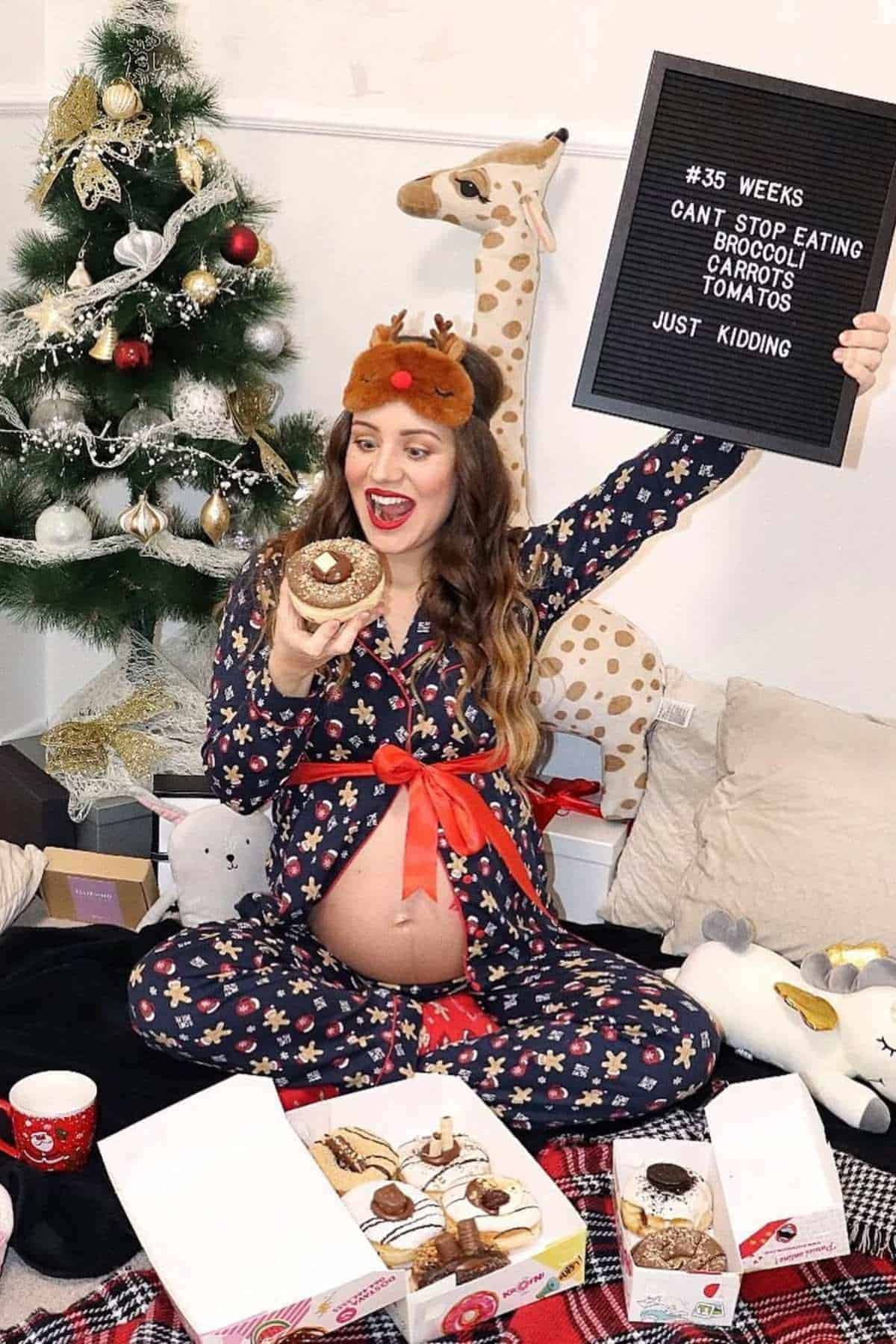 Pregnant woman in pajamas sits by a Christmas tree, holding a donut and a letter board with a humorous pregnancy craving message. Boxes of donuts and holiday decorations are around her.