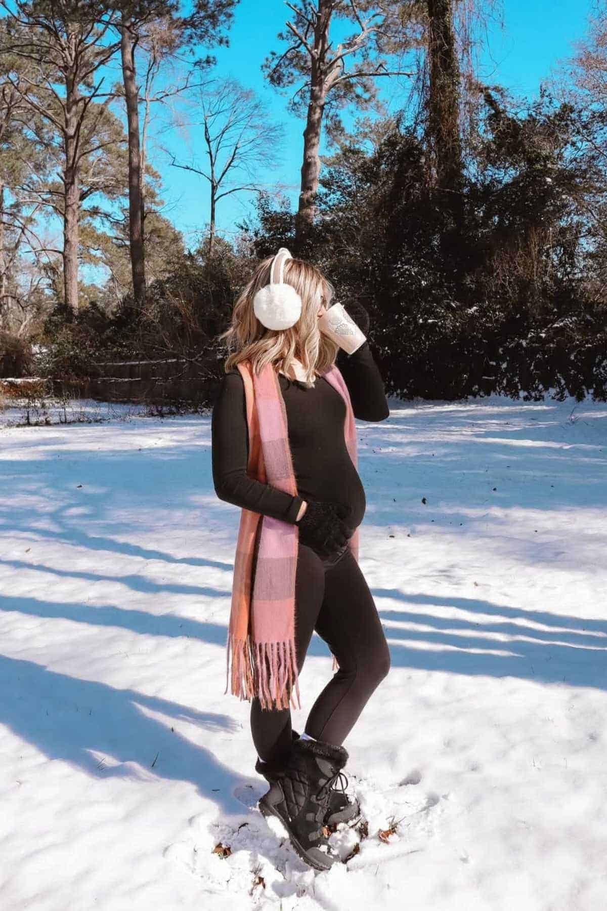A woman in winter clothing and earmuffs stands in the snow, holding a mug and wearing a pink scarf, with trees and a blue sky in the background.