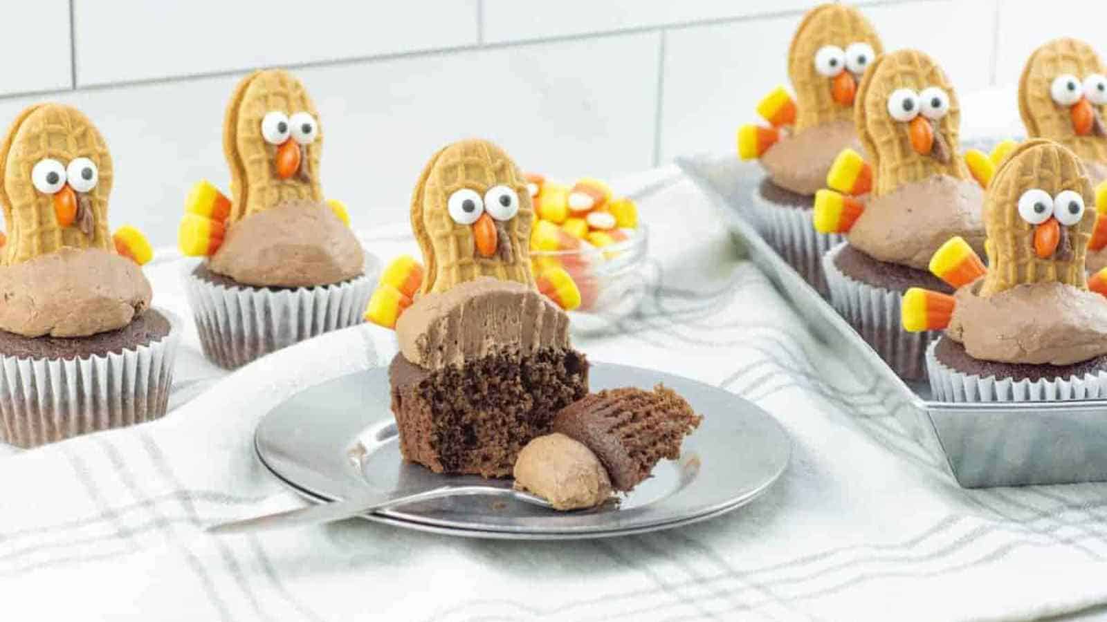 Chocolate cupcakes decorated as turkeys with cookies, candy eyes, and candy corn for feathers; one cupcake is cut open on a plate with a fork.