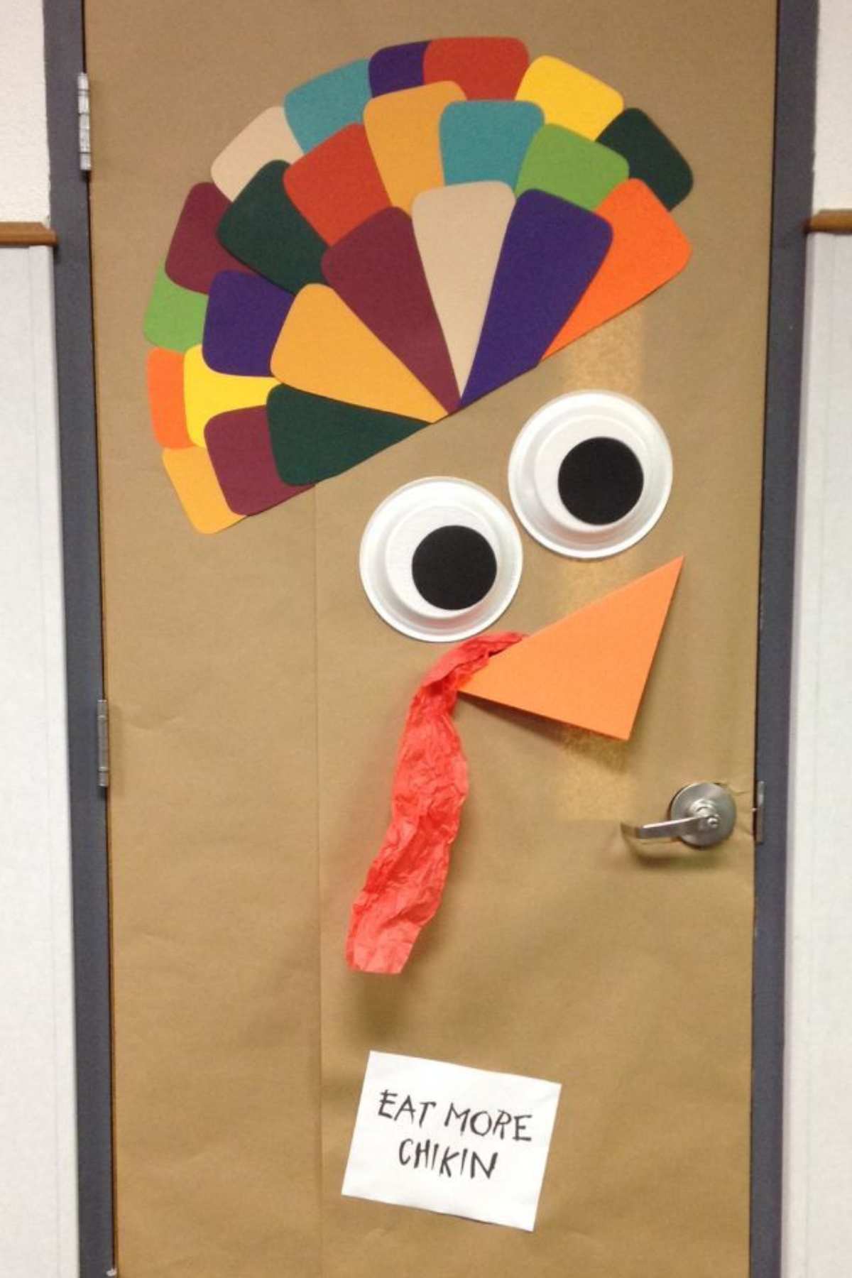 A door decorated as a turkey with colorful paper feathers, large googly eyes, an orange beak, red wattle, and a sign that says "EAT MORE CHIKIN.
