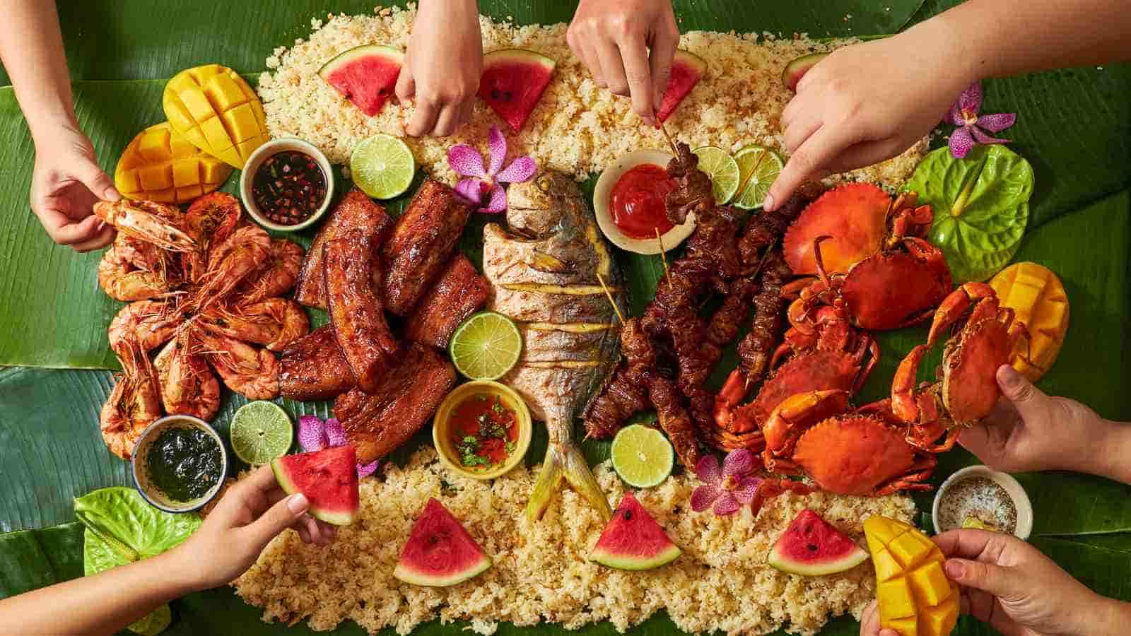 Hands reaching for various seafood, grilled meats, rice, mango, and watermelon pieces arranged on banana leaves in a communal style meal.