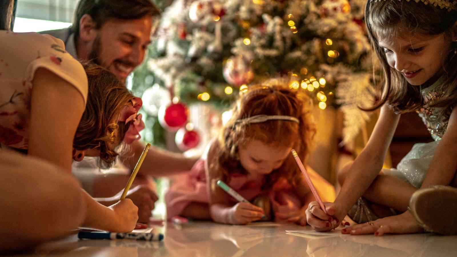 Three children draw on paper with colored markers on the floor near a decorated Christmas tree while an adult watches and smiles in the background.