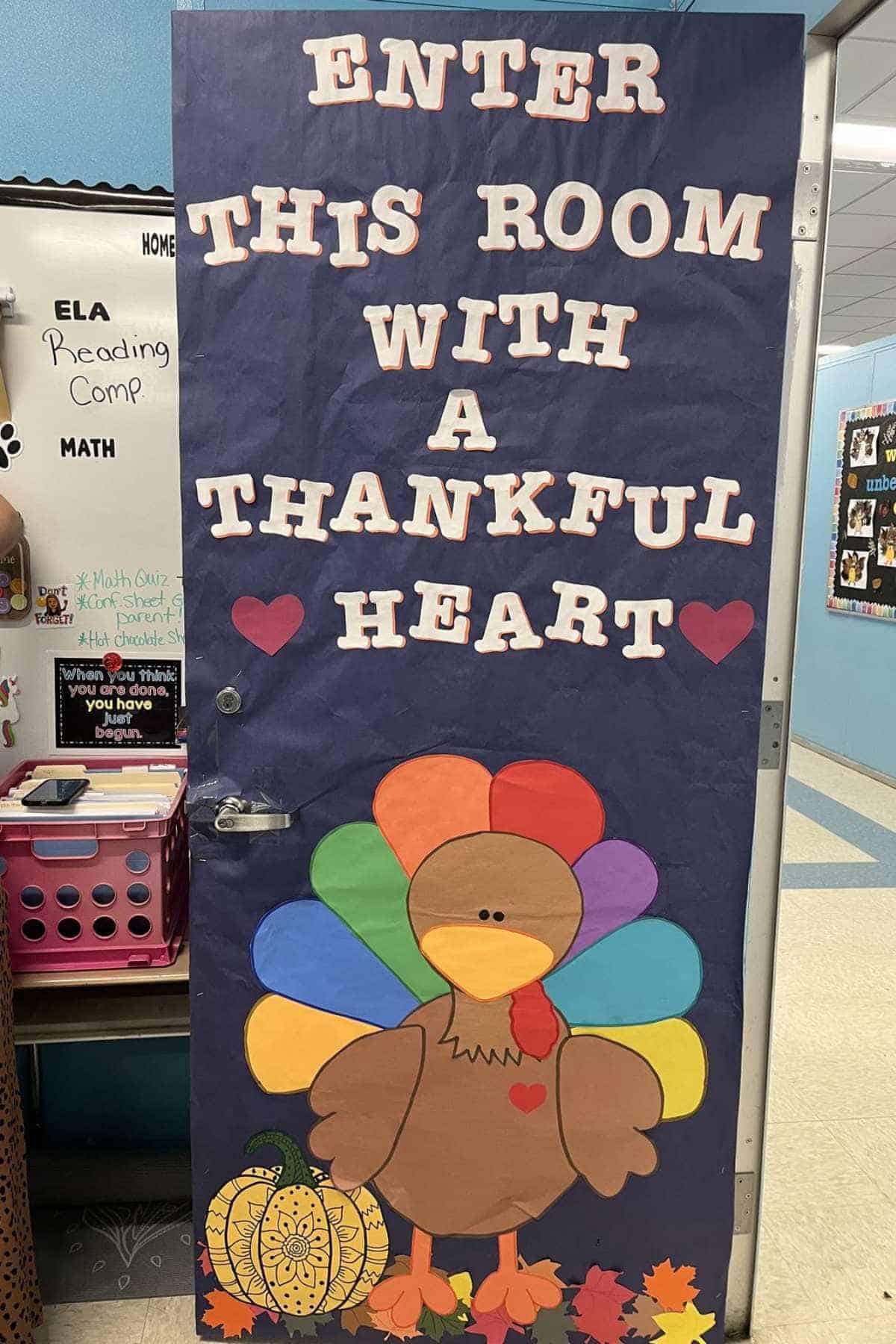 Classroom door decorated with a large turkey, colorful feathers, a pumpkin, and leaves, with the message "Enter this room with a thankful heart" in large white letters.