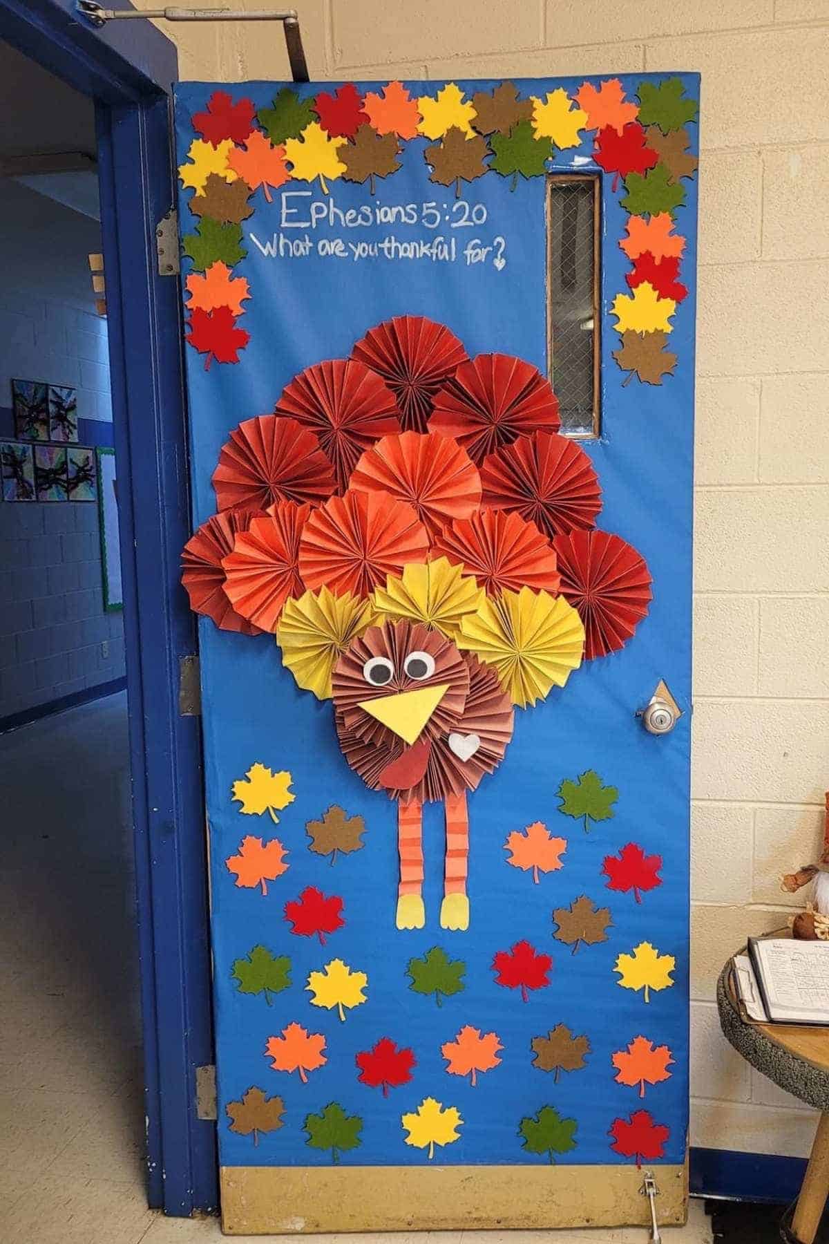 A blue door decorated with a large paper turkey and colorful paper leaves, featuring the text "Ephesians 5:20 What are you thankful for?" written in white above the turkey.