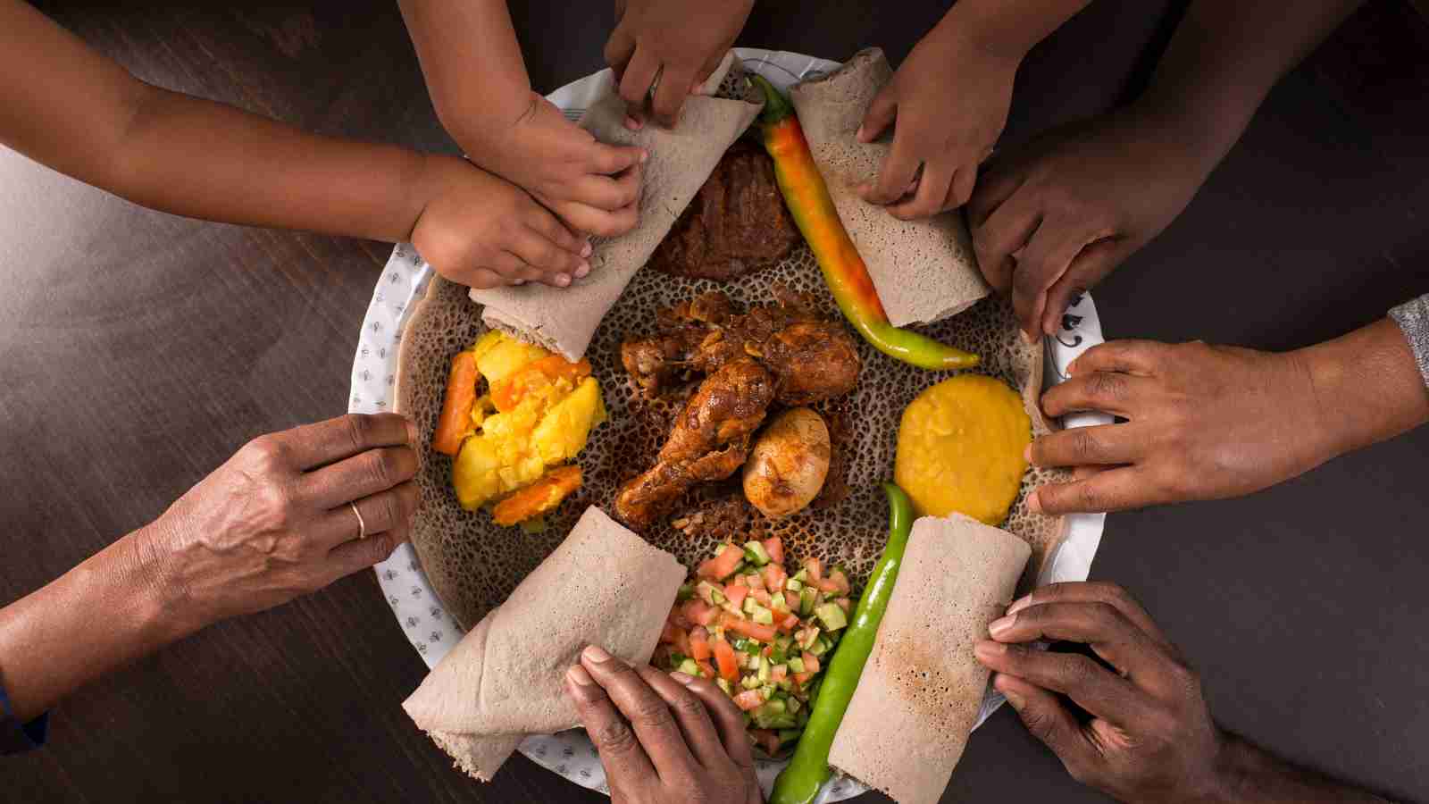 Several hands reach for pieces of injera and assorted Ethiopian dishes arranged on a large shared plate.