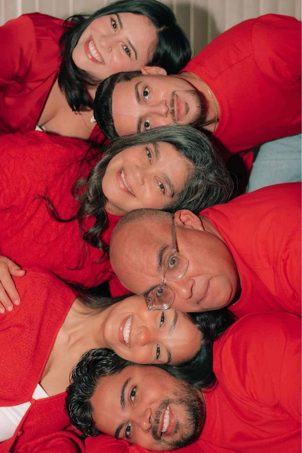 Six people, three men and three women, wearing red tops, lie close together in a face stack, smiling at the camera against a neutral background.