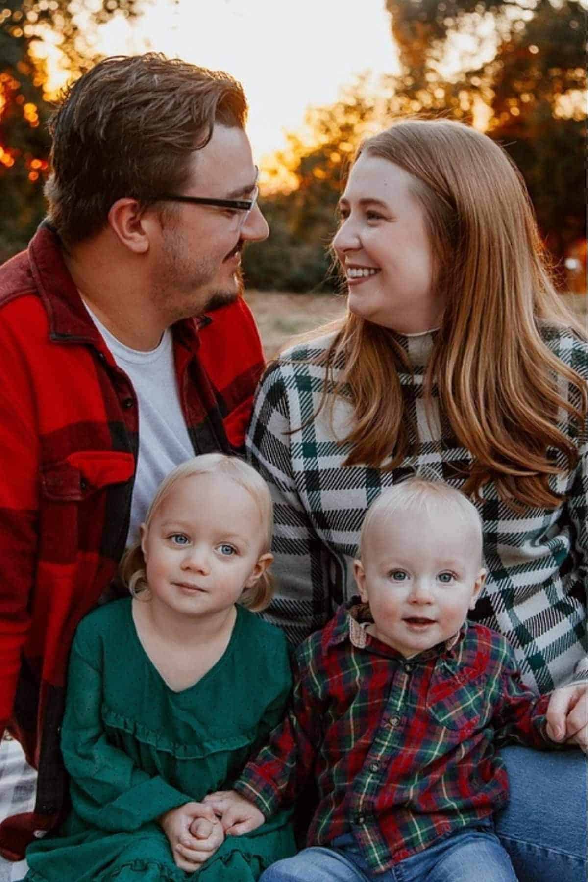 A man and woman sit outdoors at sunset with two young children, all smiling at the camera. The adults wear plaid shirts; the children wear green and plaid outfits.