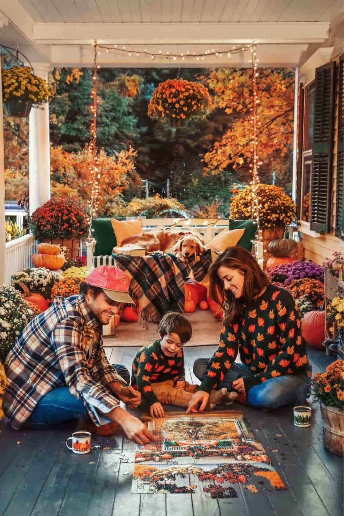 A family of three and a dog sit on a decorated porch doing a puzzle, surrounded by autumn leaves, pumpkins, flowers, and string lights.