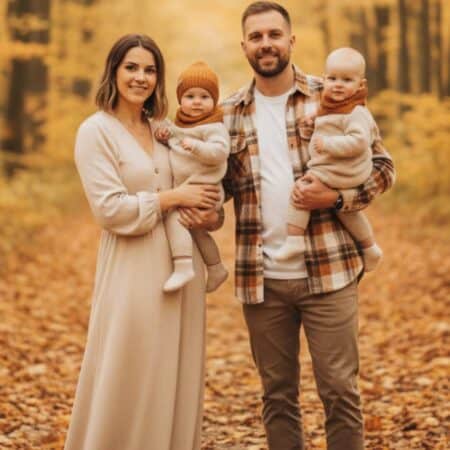 A man and woman stand in a forest with autumn leaves, each holding a baby dressed in warm clothing.
