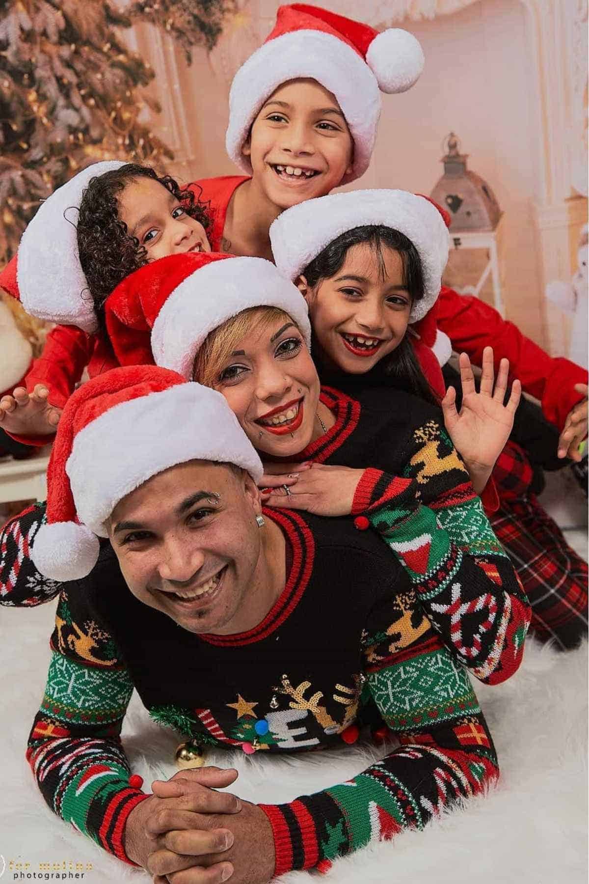 Five people in Christmas sweaters and Santa hats are stacked in a playful pose, smiling in front of a festive holiday background.