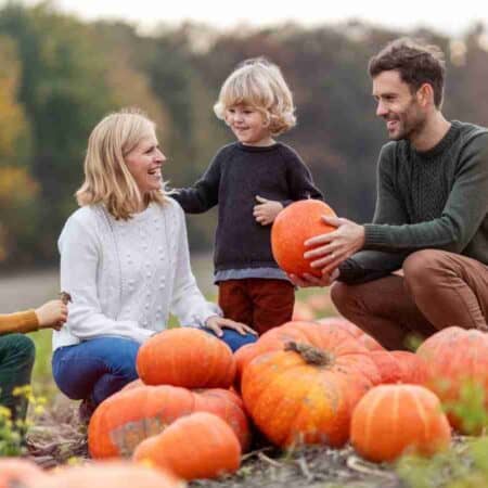A family of four sits and kneels around large pumpkins in a pumpkin patch on a cloudy day, surrounded by fields and trees.