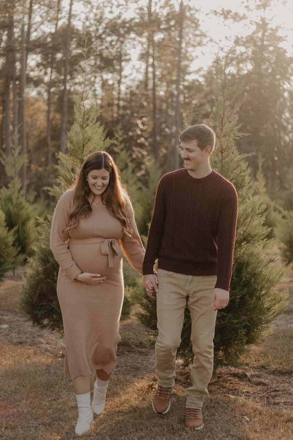 A pregnant woman and a man walk hand in hand outdoors among small pine trees, both smiling and dressed in casual, neutral-colored clothes.