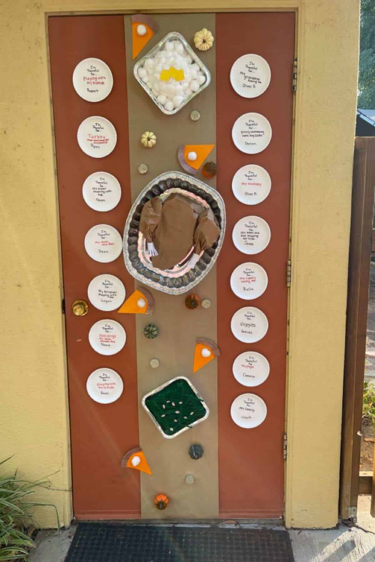 A door decorated with paper plates listing Thanksgiving foods, small gourds, and representations of pie, mashed potatoes, and a turkey.
