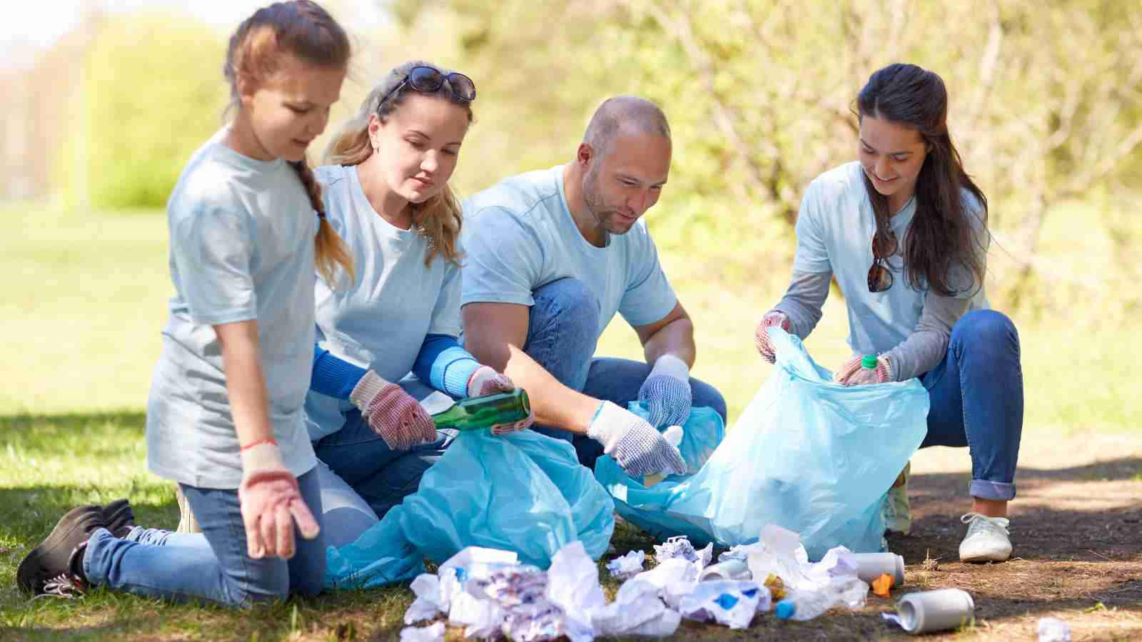 Four people wearing gloves pick up litter and place it into blue trash bags in a grassy outdoor area.