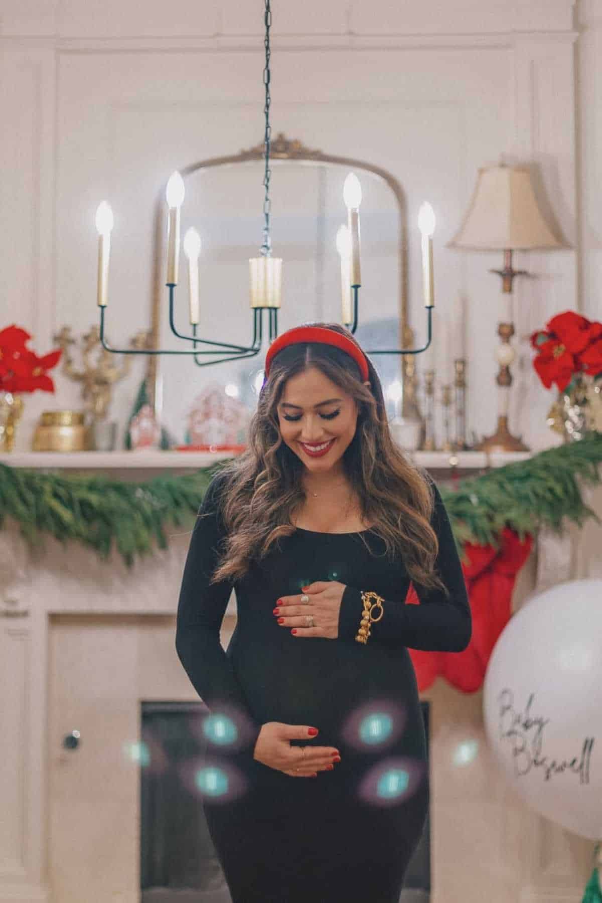 A woman in a black dress stands indoors, smiling and holding her pregnant belly. The room is decorated with holiday greenery, red bows, and a balloon that says “Baby Bloomwell.”.