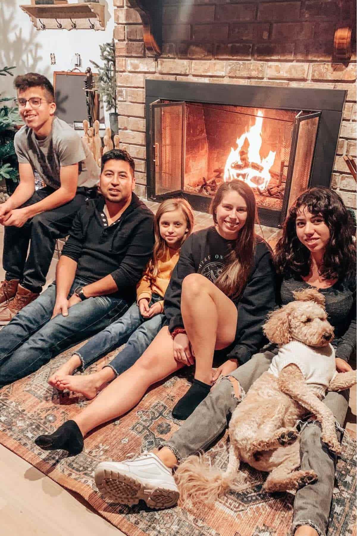 Five people and a dog sit on a rug in front of a lit fireplace, smiling at the camera in a cozy indoor setting.