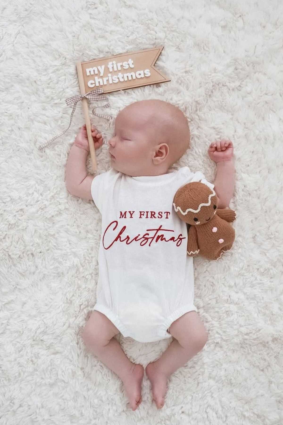 A baby sleeps on a white rug wearing a "My First Christmas" onesie, holding a plush gingerbread toy, with a "my first Christmas" sign nearby.