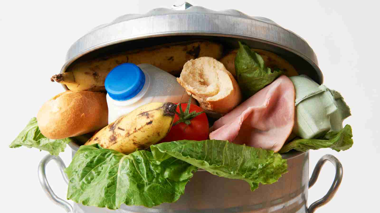 A metal trash can filled with discarded food items, including bananas, bread, lettuce, a tomato, ham, a plastic bottle, and an egg carton.