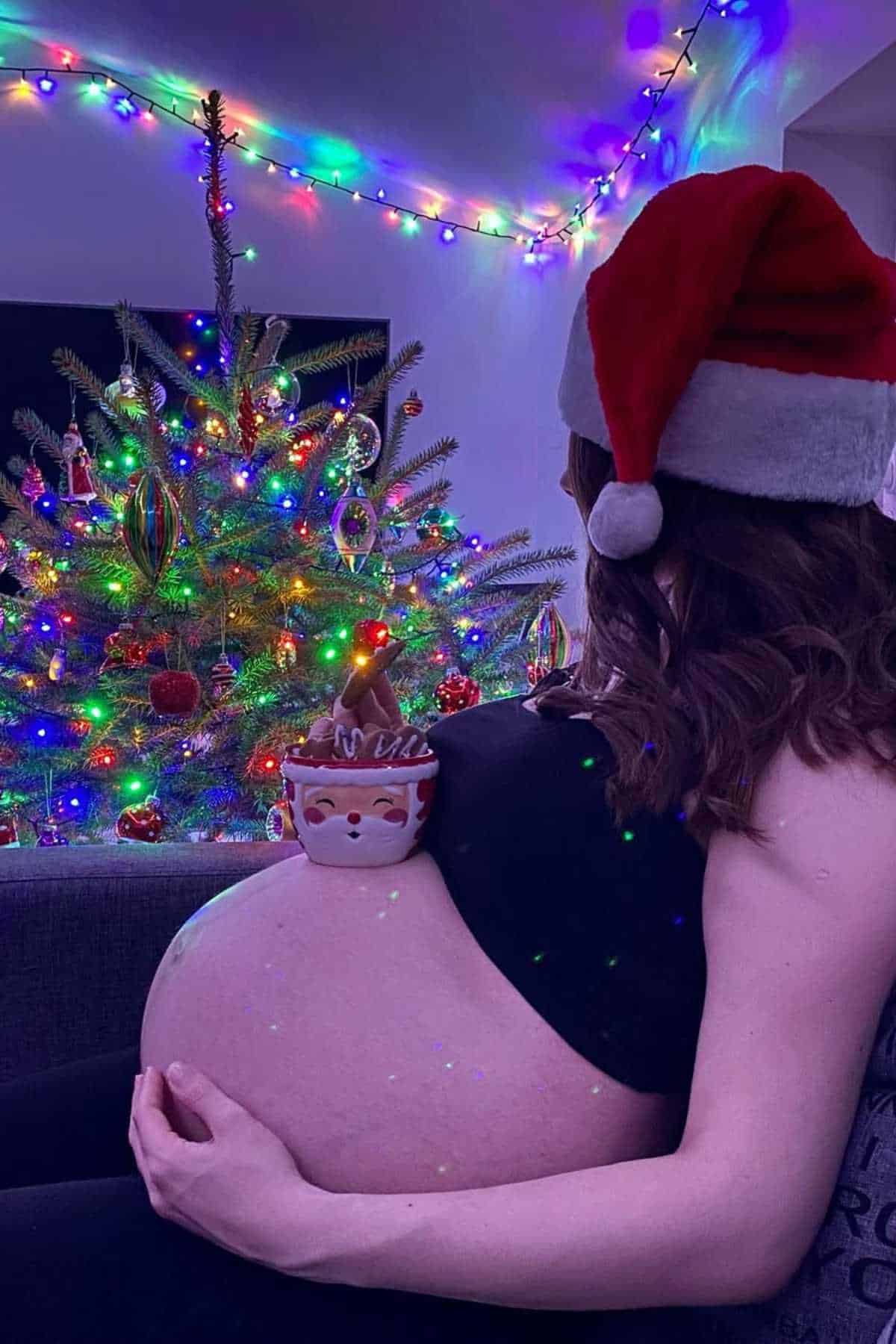 A pregnant person wearing a Santa hat balances a holiday mug on their belly, sitting near a decorated Christmas tree with colorful lights.