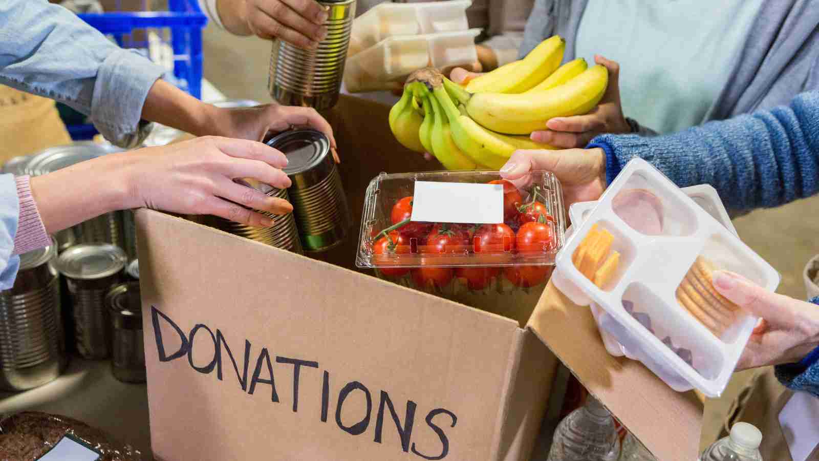 Several hands place canned goods, bananas, tomatoes, and packaged food into a box labeled "Donations.