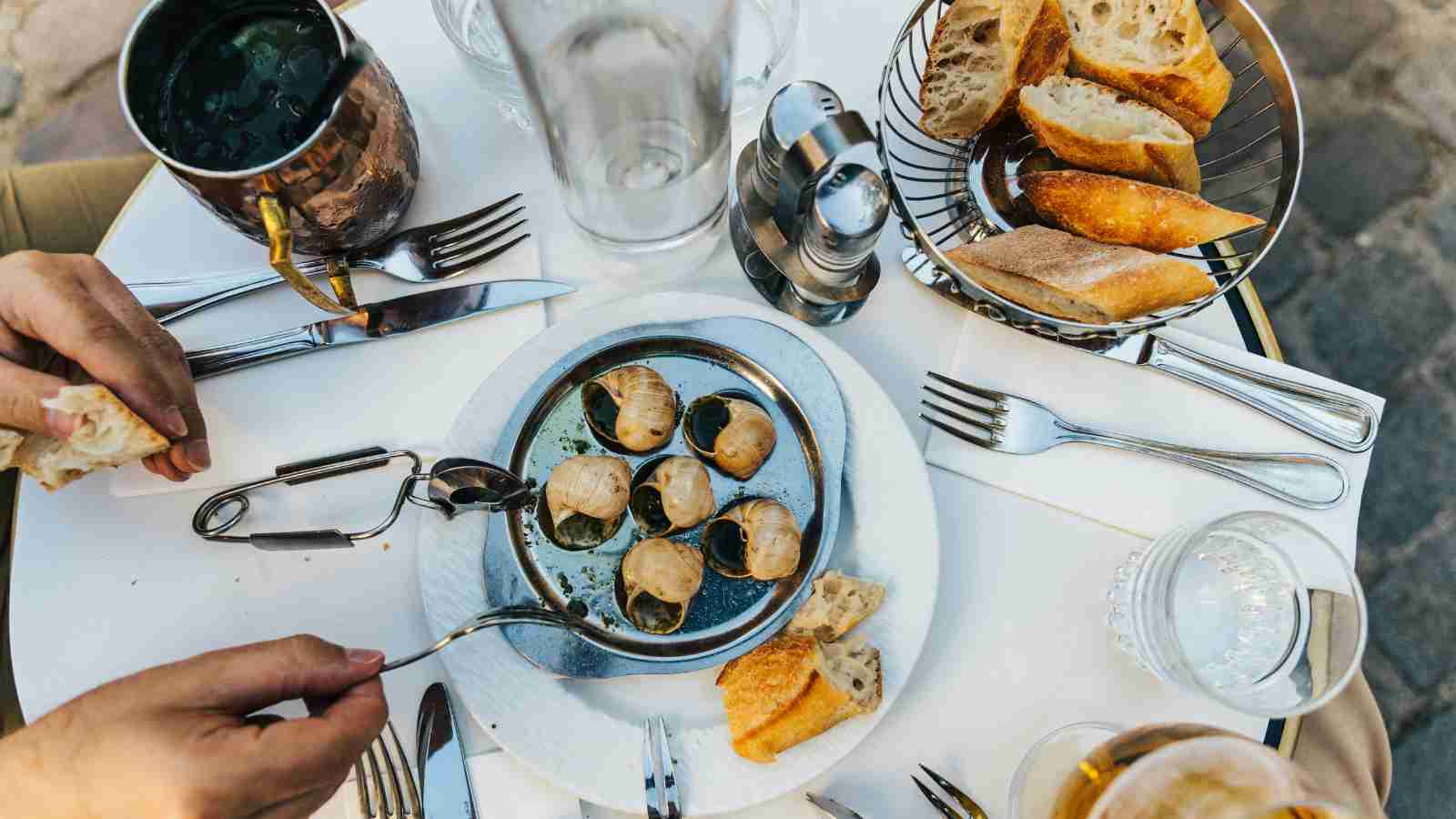 A table set with escargot, sliced baguette, utensils, a glass of water, a drink in a metal cup, and salt and pepper shakers, viewed from above.
