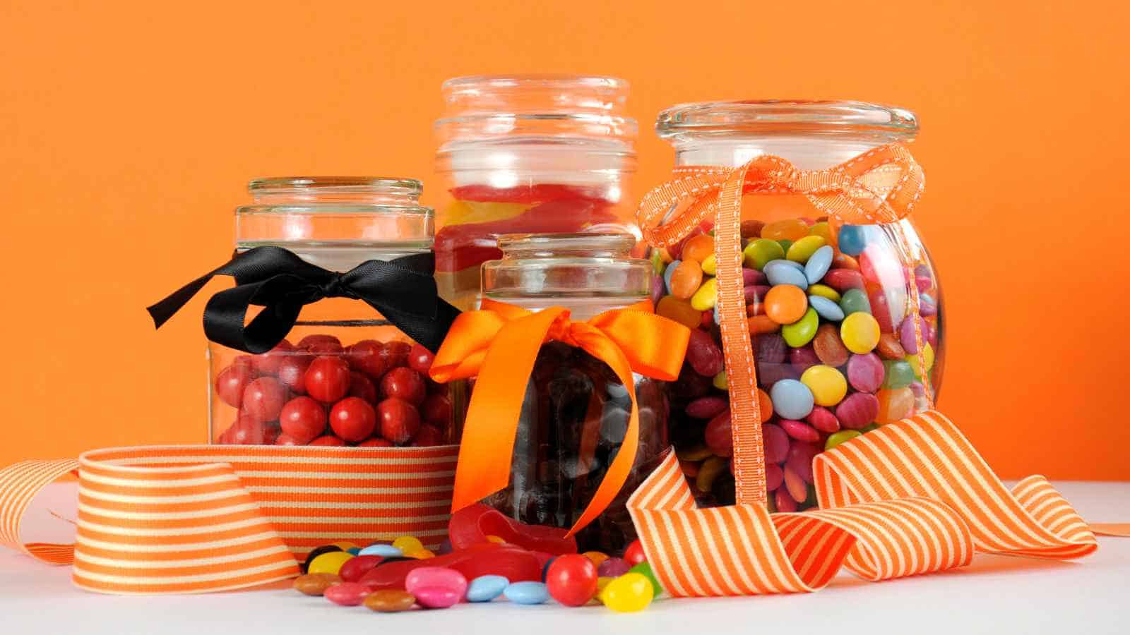 Four glass jars filled with colorful candies and tied with black or orange ribbons, surrounded by loose candies and orange-striped ribbon, set against an orange background.