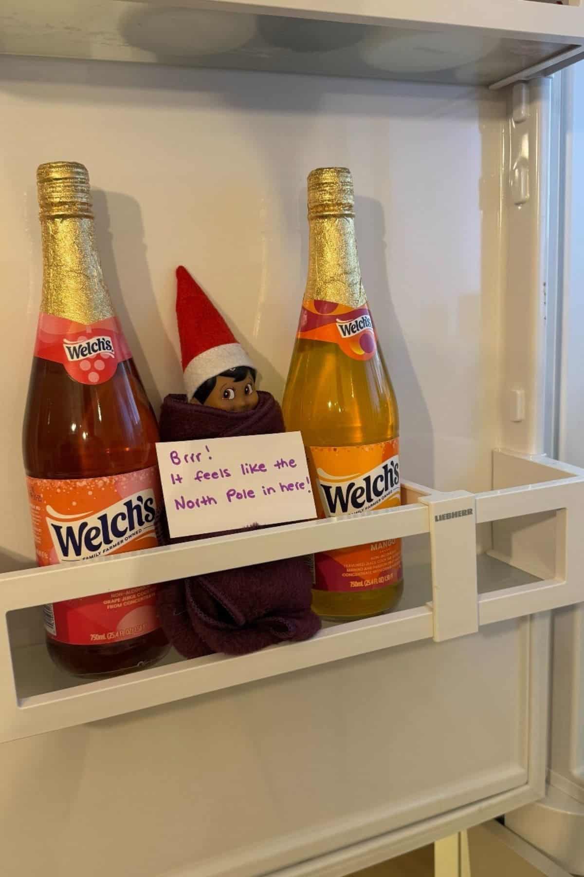 A small elf doll wrapped in a cloth sits between two bottles of Welch's juice in a fridge door with a note reading, "Brrr! It feels like the North Pole in here.