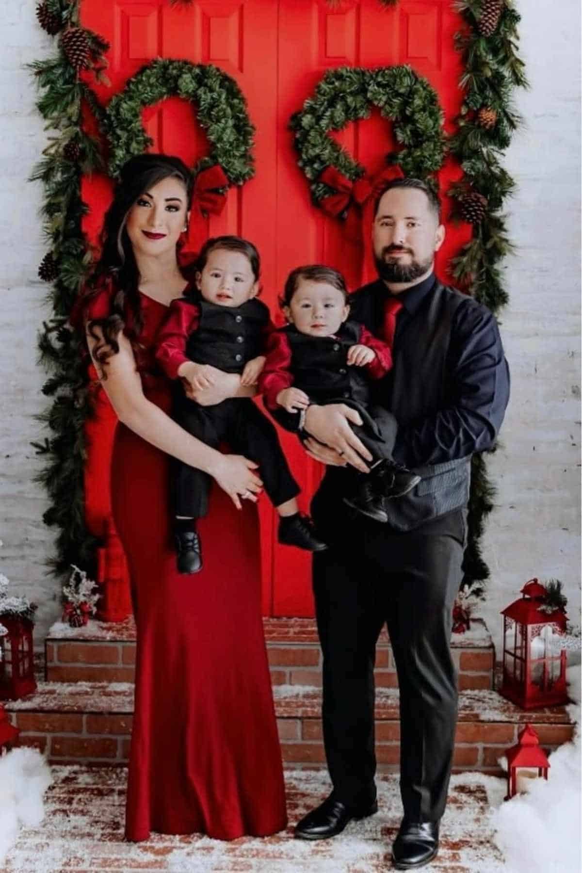 A family of four poses in front of a red door decorated with wreaths and greenery, all dressed in coordinated black and red outfits.