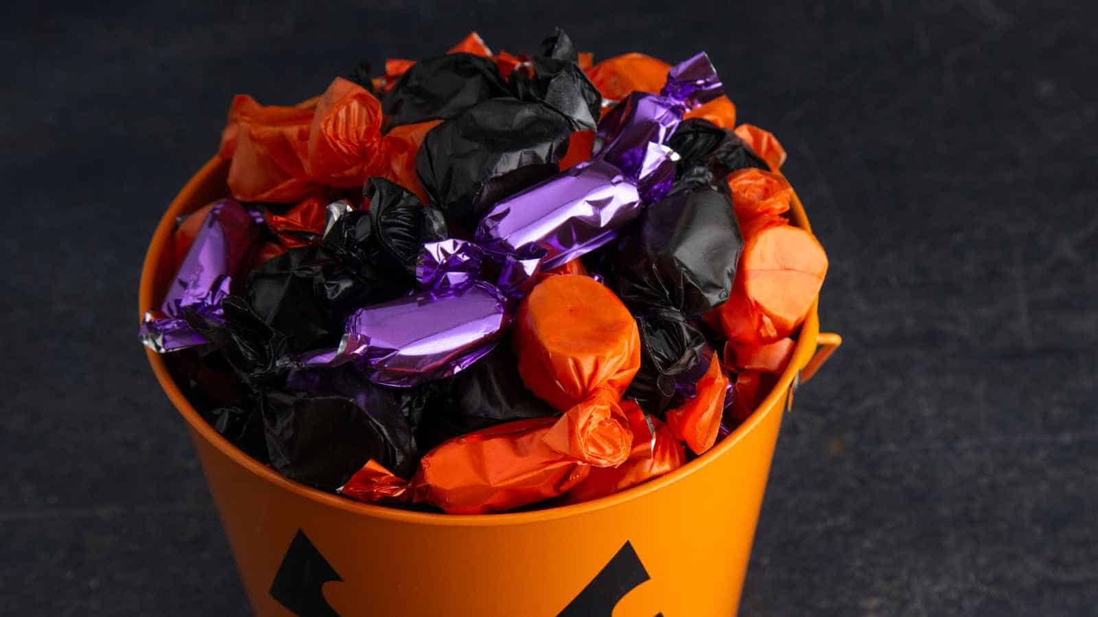 An orange bucket with a jack-o’-lantern face is filled with assorted candies wrapped in black, orange, and purple wrappers.