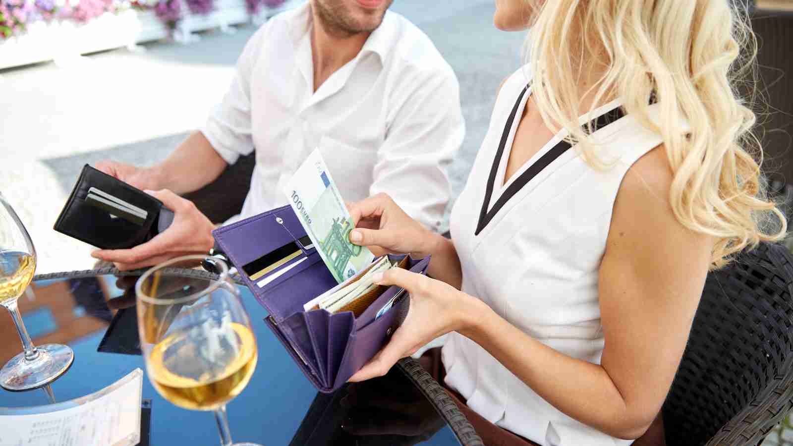 Two people sit at an outdoor table with drinks. The woman holds euro banknotes and an open wallet, while the man opens his wallet.
