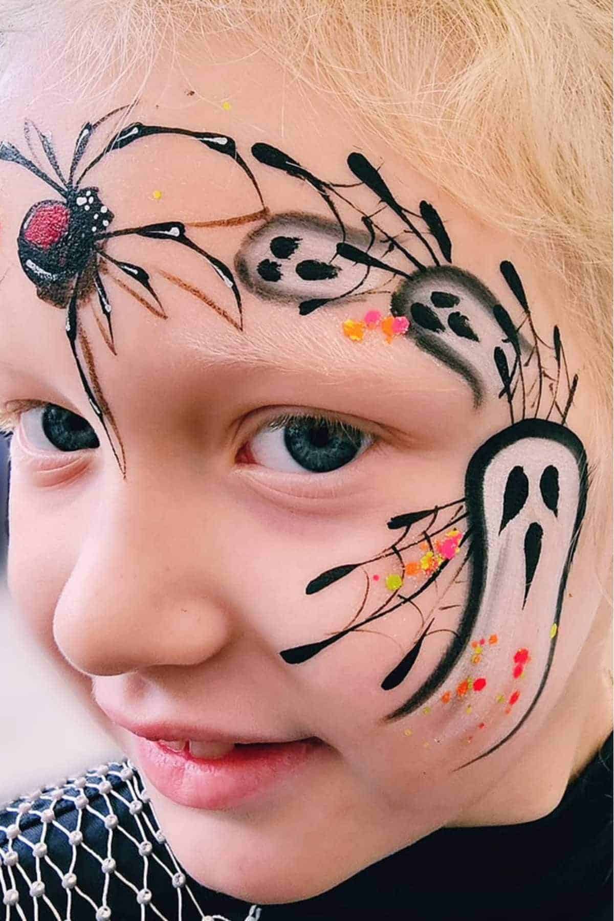 A child with blonde hair has face paint of black spiders, webs, and ghost faces with red and yellow dots on one side of their face.