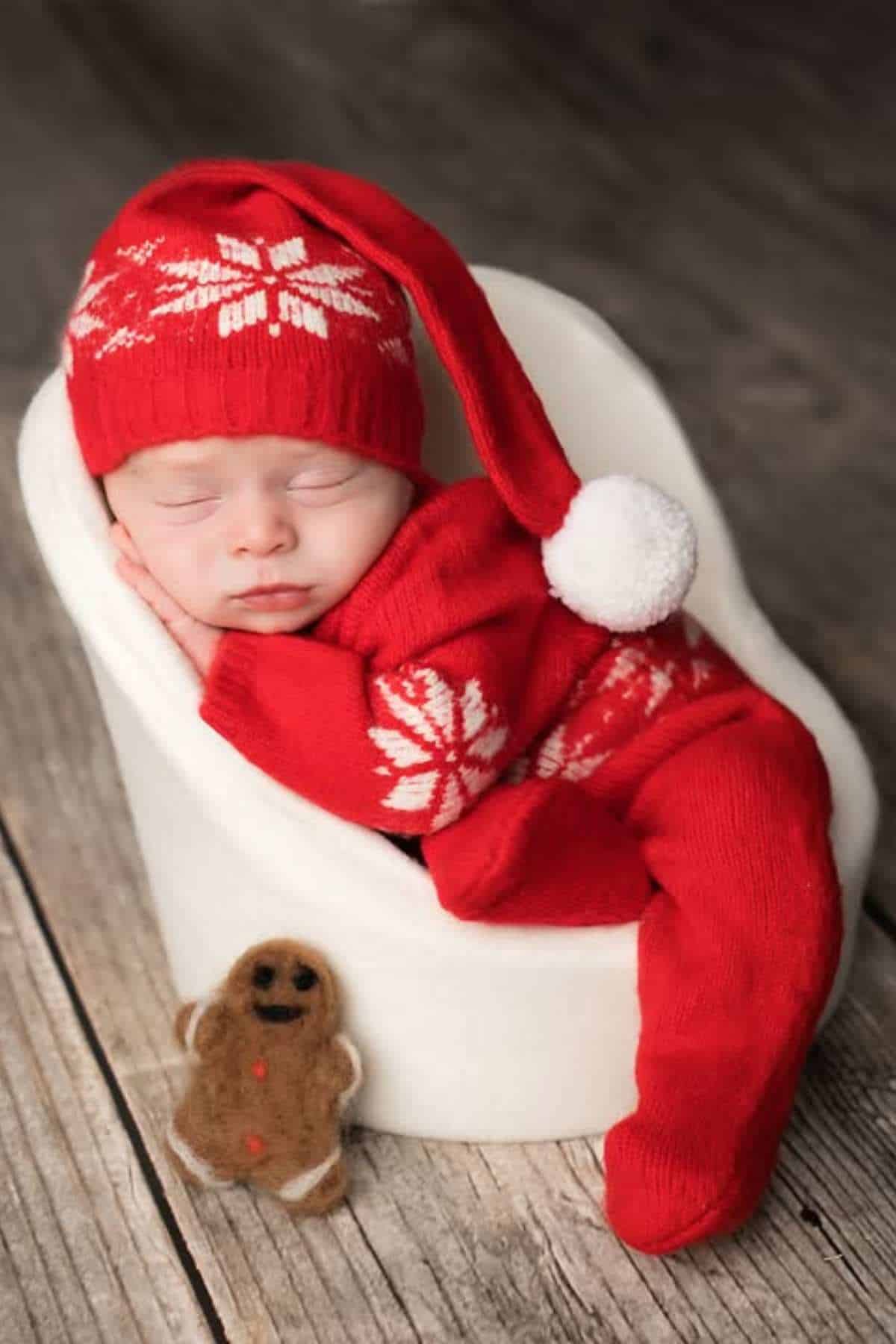 A baby wearing a red knit hat and outfit with white snowflake patterns sleeps in a white chair next to a small gingerbread man toy on a wooden floor.